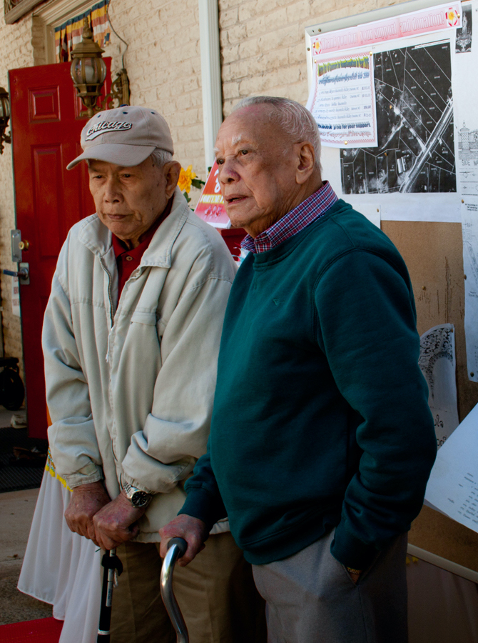 Two elderly men stand next to each other leaning on mobility aids.
