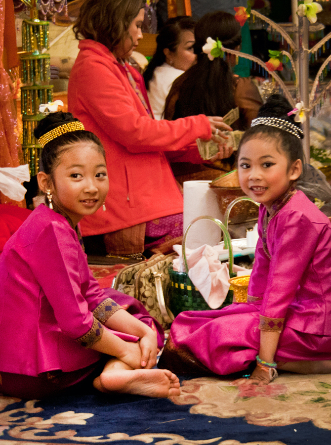 Two young girls wearing bright pink jackets and skirts adorned with intricate embroidery. They are seated on the floor while a woman behind them counts cash.