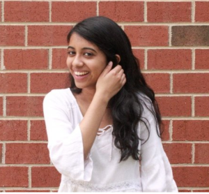 Maydha Devarajan, the founder of Claiming Carolina, smiles into the camera in front of a brick wall. She is wearing a white shirt.